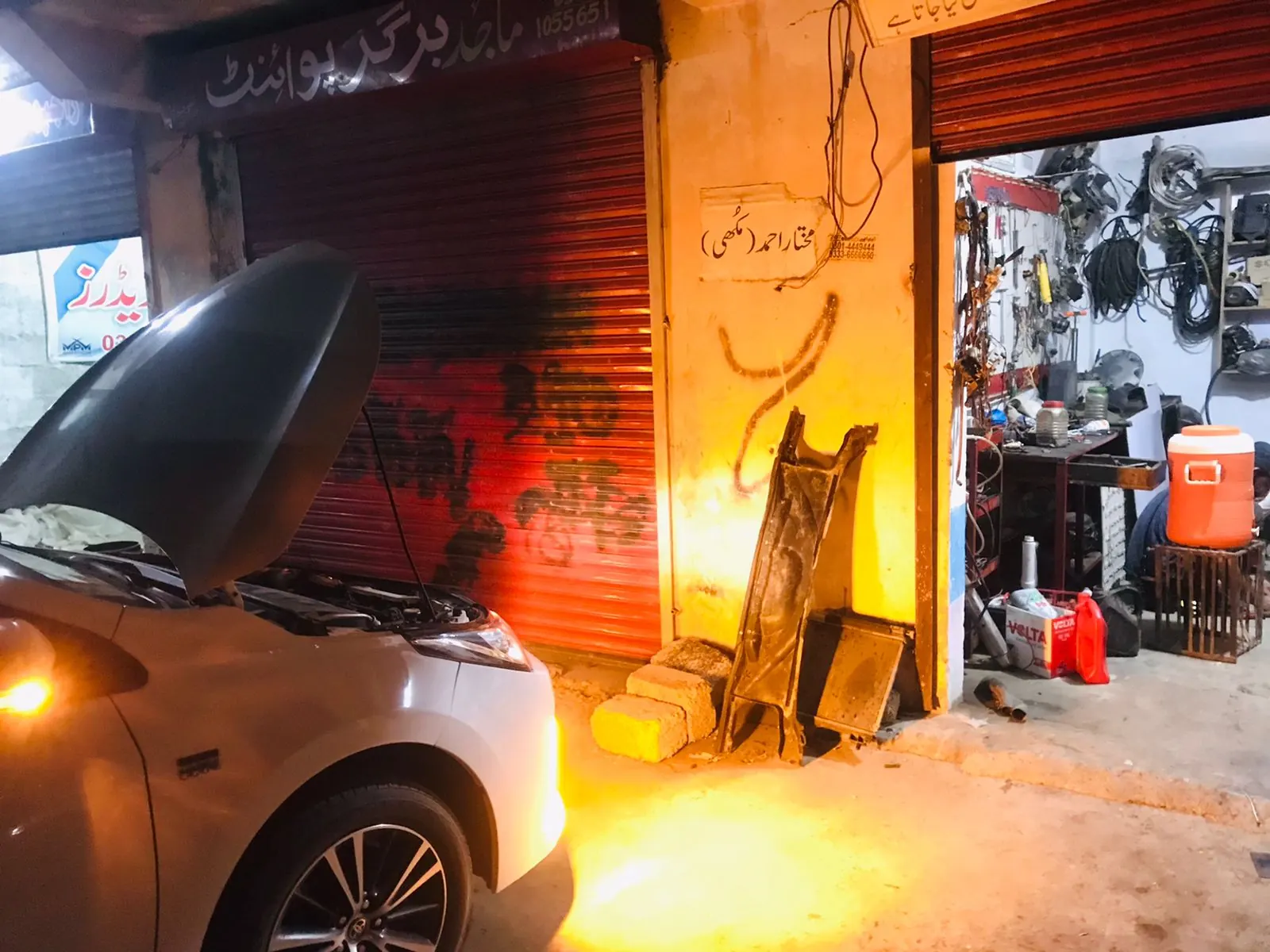 A car with its bonnet up in the workshop bay at night, tools and wiring on the wall